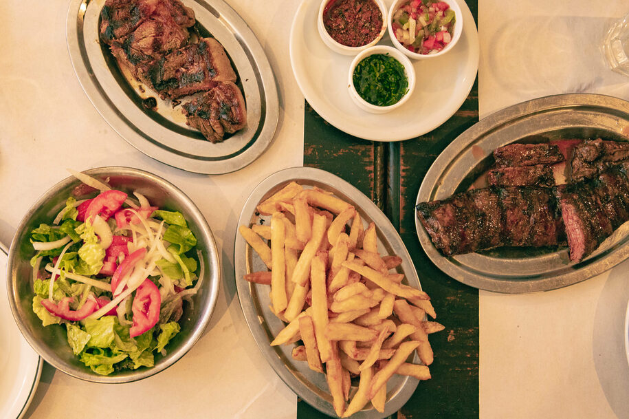 Steak, fries, salad and three small bowls containing chimichurri, salsa criolla and provenzal sauce. Steak is an integral part of Argentina's Food Culture and the most internationally recognized Argentine food.