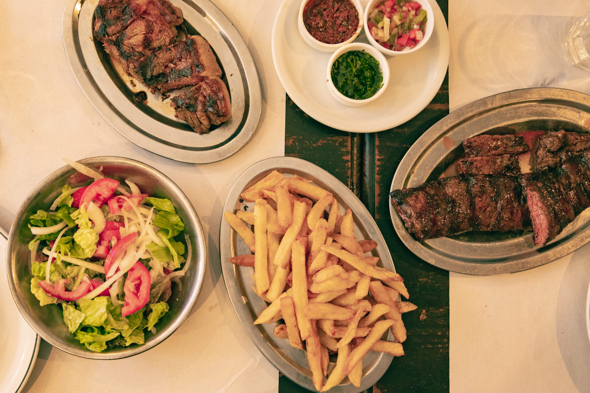 Steak, fries, salad and three small bowls containing chimichurri, salsa criolla and provenzal sauce. Steak is an integral part of Argentina's Food Culture and the most internationally recognized Argentine food.