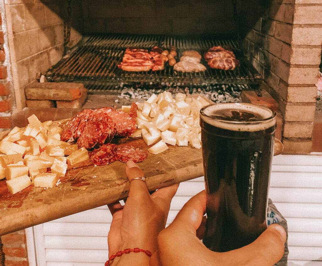 A wooden cutting board with sliced salami and cheese with a glass of fernet cocktail (fernet with coke). There is a grill on the background, with several cuts of meat on it.