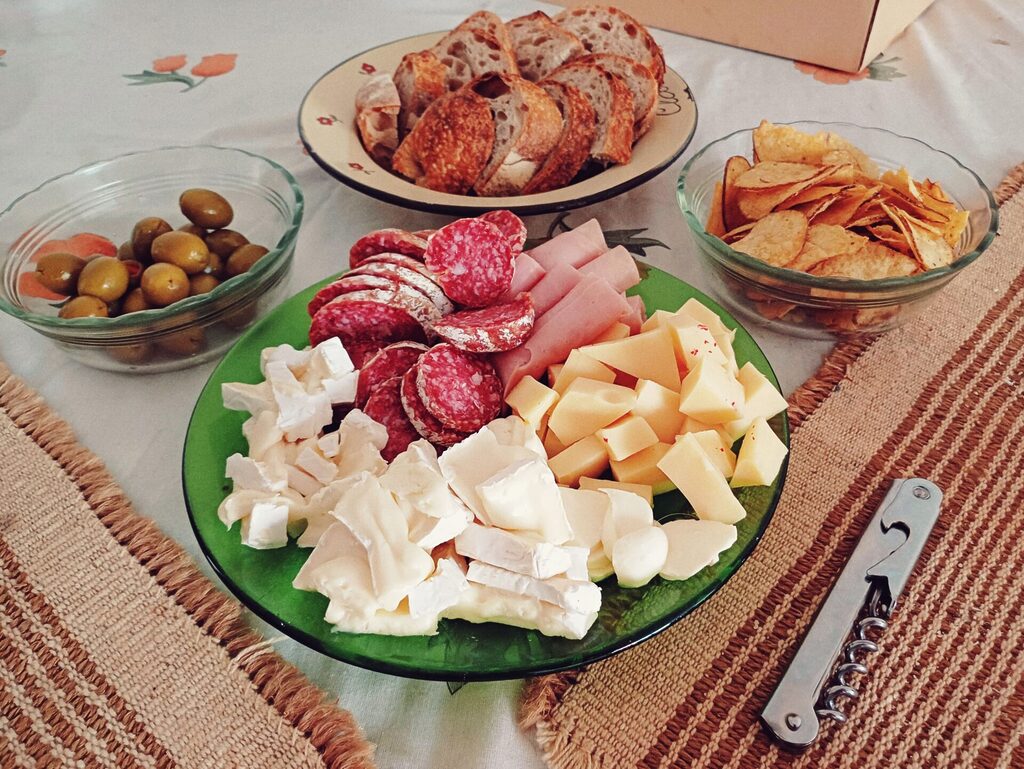 An Argentina picada: on a green plate, salami, brie cheese, ham slices and holanda cheese are served. There's a small glass bowl with olives, a plate with slices of bread and another glass bowl with crisps.