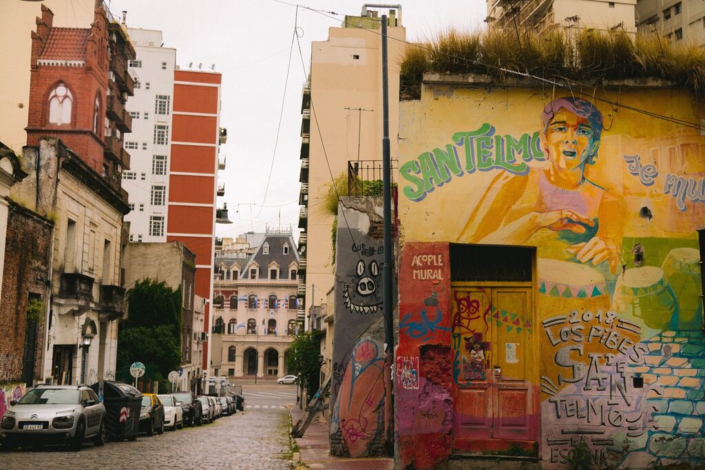 A picture of a street in San Telmo. A cobbled street leads to an imposing building. On the foreground, a house features a colorful mural with some text and phrases about the neighborhood and its culture.