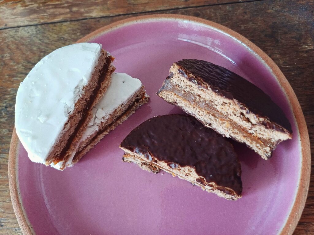 Two alfajores cut in half served on a pink plate. The alfajor on the left has a sugar glaze coating and the alfajor on the right has a chocolate coating.