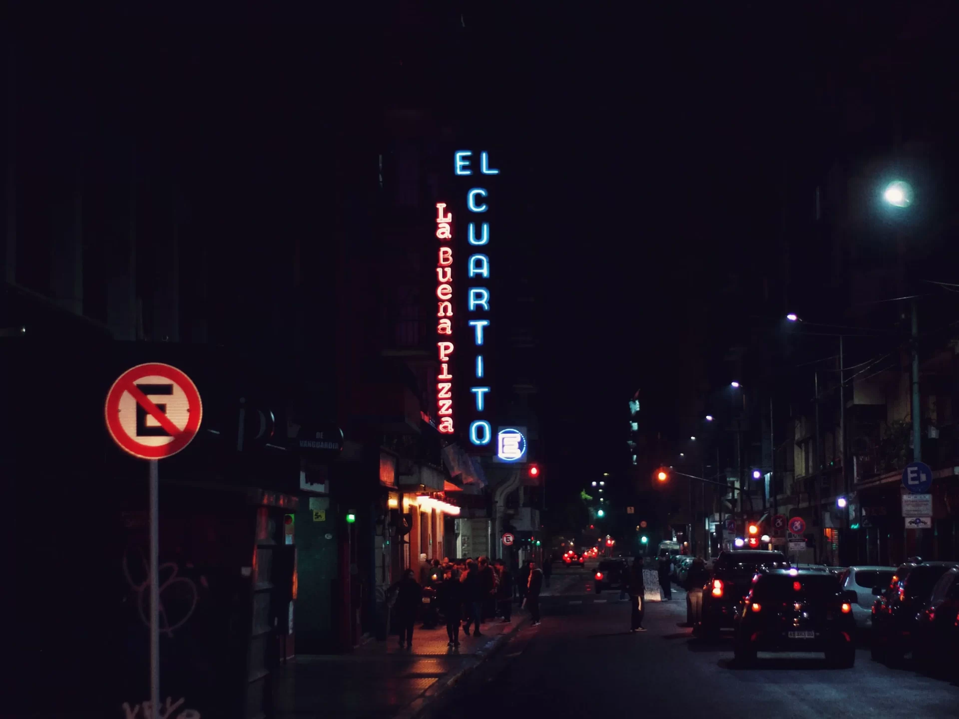 Neon sign with the words "El Cuartito, la buena pizza" featured to the side of a pizza store of the same name. The picture was taken during night time. El Cuartito's is for many the best pizza in Buenos Aires.