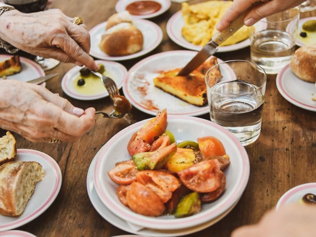 A table is set, with a plate of tomatoes, a plate of fainá and a plate of tortilla. People are cutting and serving the food.